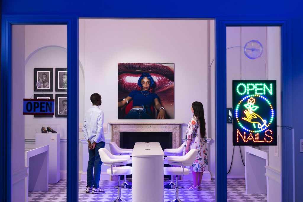 View of a temporarily installed nail shop with a neon 'Open Nails' sign featuring a hand holding a rose. Inside are two people looking at photographs of a model.