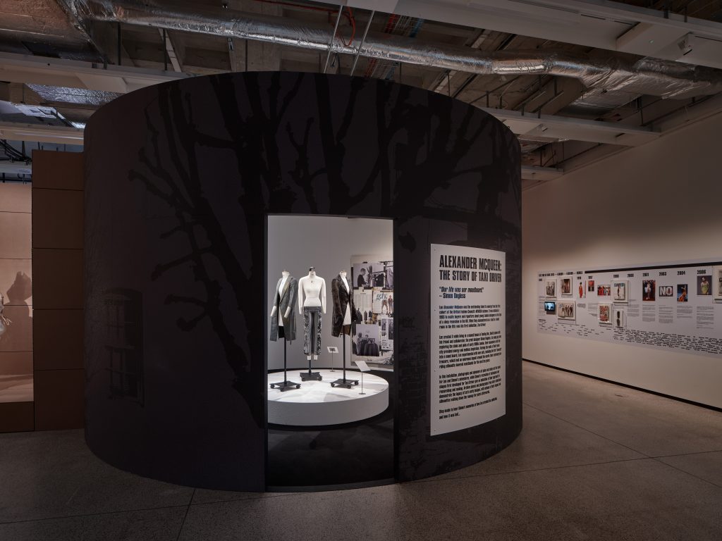 View of a grey circular installation within a gallery, featuring a label entitled 'Alexander McQueen: The Story of Taxi Driver'. Inside are three monochrome outfits on a white circular plinth.