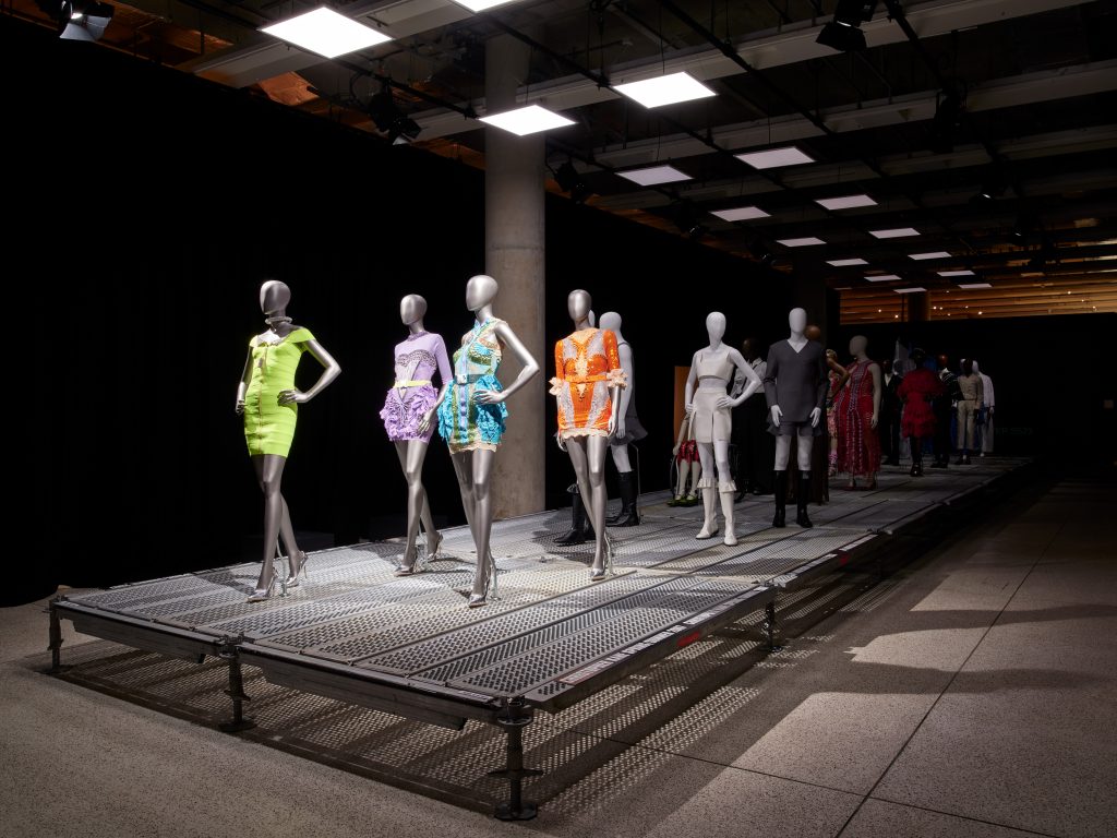 Silver mannequins stand on a grated metal catwalk, each posing wearing different clothes underneath square lighting.