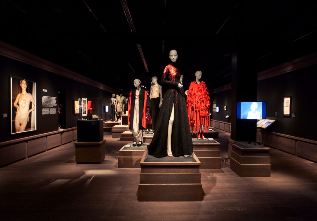 Gallery view showing a group of mannequins wearing a variety of black, red and white clothes, mostly dresses, on grey wooden plinths. Photographs and films are shown on the surrounding walls.