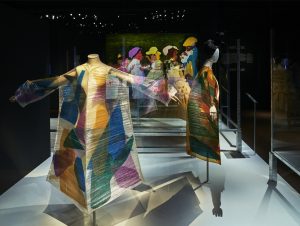 Gallery view showing two mannequins with outstretched arms wearing transluscent white dresses overlaid with colourful geometric shapes overlaid. In the background is an illustration of smiling people.