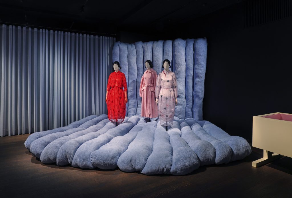 Three mannequins, each with a matching wig and Peter Pan collar, stand on a blue wide-ribbed velvet structure. From left to right, the mannequins wear a red patterned dress with long sleeves, a pink robed coat, and pale purple embroidered dress.