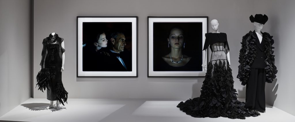 Three white mannequins wear black mannequins in front of two dark portrait photographs: a a couple and a woman.