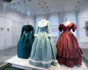 A trio of female mannequins wearing historical full length gowns, in hues of teal blue, light blue with a white fur trim and red with ruffles.