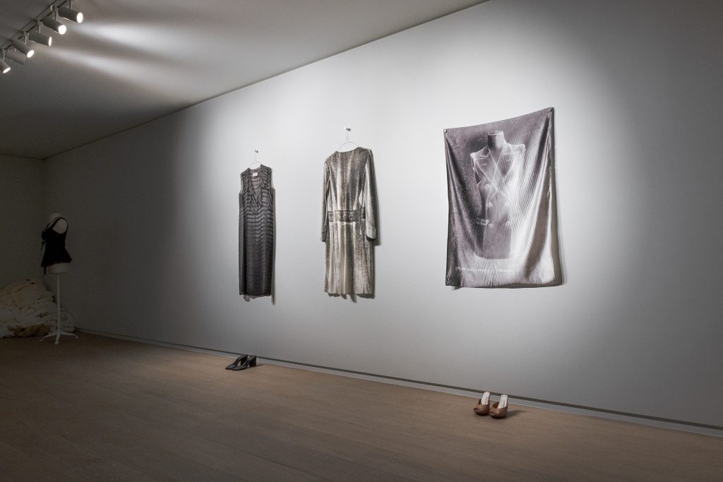 exhibition display of grey garments on gallery wall and shoes on floor
