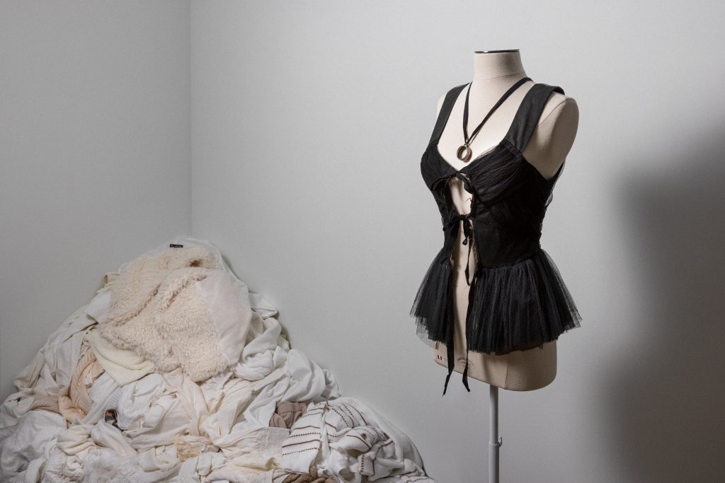 Exhibition display of mannequin dressed in black basque with pile of textiles in corner