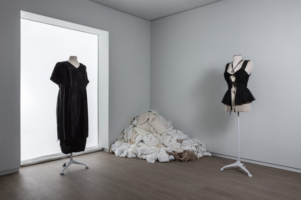 Exhibition display of mannequin dressed in black dress and black basque with pile of textiles in corner