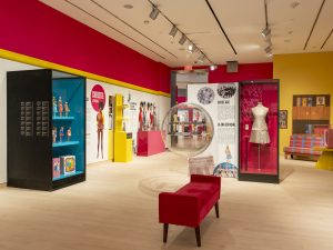 Installation of a pink Barbie dress, display cabinets of Barbie dolls in multicolours.