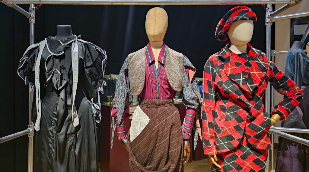 A trio of mannequins on display. Left is wearing a black dress, the middle one is wearing plaid trousers, shirt and jacket, and the right mannequin is wearing a red and black outfit.