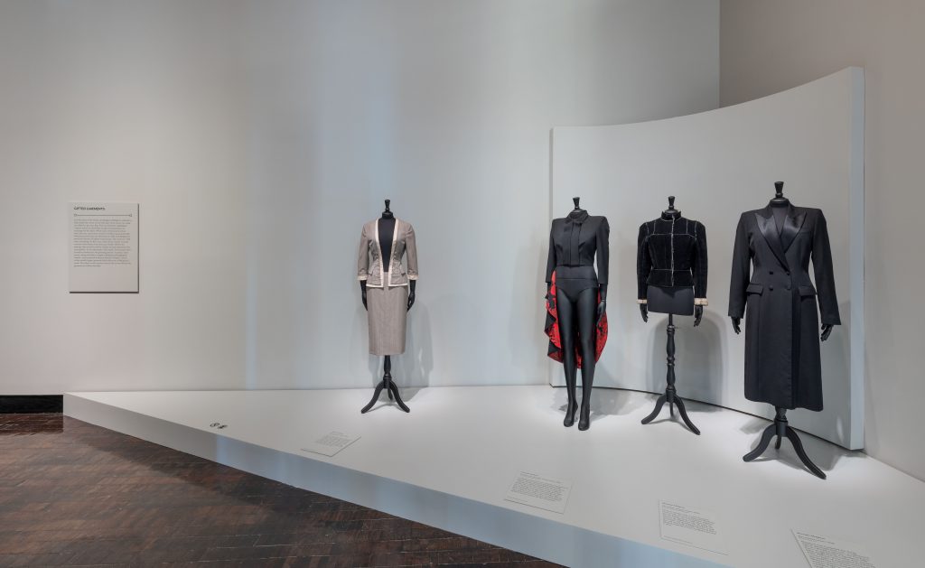 A display of 4 mannequins; right side a female mannequin wears a black trench coat, middle mannequin wears a black jacket, next mannequin is wearing a black and red piece. Lastly, the mannequin to the far left is wearing a beige top and skirt.