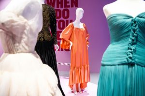 Three female mannequins. Middle mannequin is wearing a bright orange dress. Mannequin to the left is wearing a white dress and the mannequin to the right is wearing a blue dress.