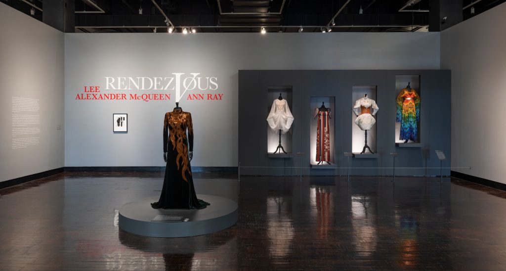 A female mannequin wearing a long black dress with gold embellishments. to the right off the image our a collection of more female mannequins wearing colourful dresses in hues of white, orange, blue and green.