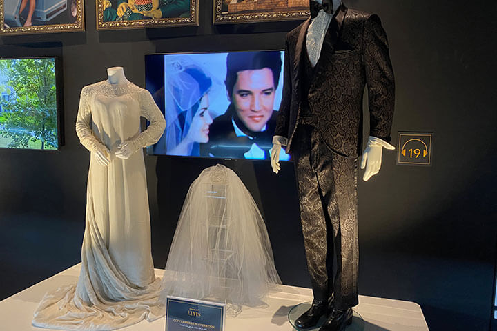 A display of Elvis and Priscilla's wedding garments. On display is a 1960's style white wedding dress complete with veil on a separate display stand. A male tuxedo suit, styled with a white suit and bow tie.