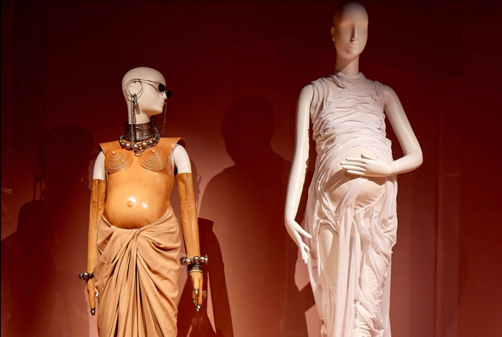 Two 'pregnant' female mannequins on display. Left: Mannequin is wearing a tan armour like top with a brown wrap skirt. Right: with two piece ruffled top and skirt.