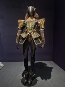 Costume for Catherine of Aragon from ‘Six: The Musical’, 2020, designed by Gabriella Slade. Displayed in ‘Six Lives: The Stories of Henry VIlI’s Queens’ at The National Portrait Gallery, London. The costume is mostly black and gold. Puff shoulders of gold and black tights, with black shoes.