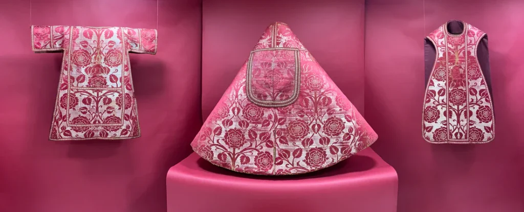 A display of red and gold garments. Left: a T-shaped garment hung up on a red backdrop. Centre: a cone shaped red skirt. Right: a red and gold patterned dress.