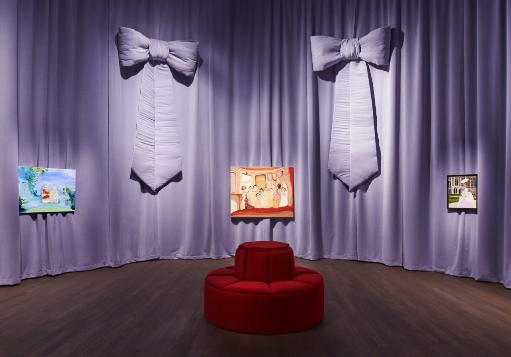 A lilac backdrop with two bows mounted up high. A red plush chair is placed in the middle of the gallery. There are colourful artworks along the walls.