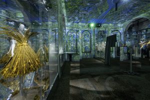 installation shot of mannequin with dress made of yellow and black feathers with image reflected behind