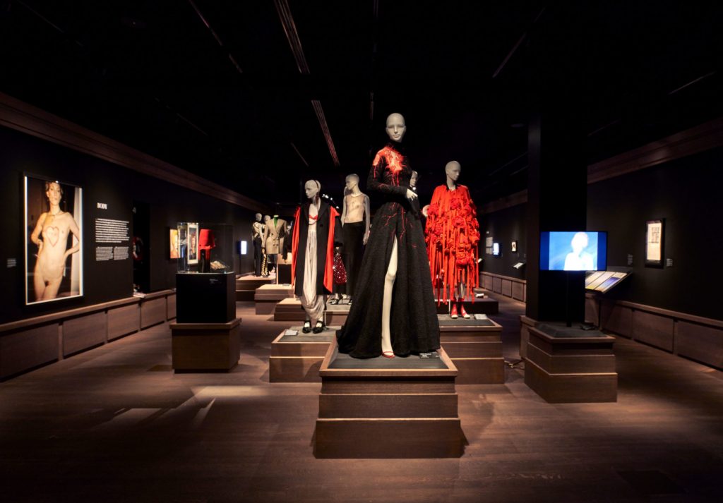 Four female mannequins: Centre mannequin in all black, full length outfit. Background mannequins wearing combinations of black and red garments.