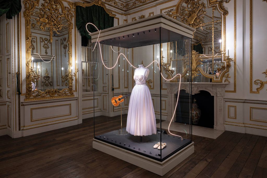 A white full length dress displayed in a vitrine alongside a guitar and book.