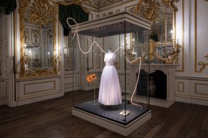 A white full length dress displayed in a vitrine alongside a guitar and book.