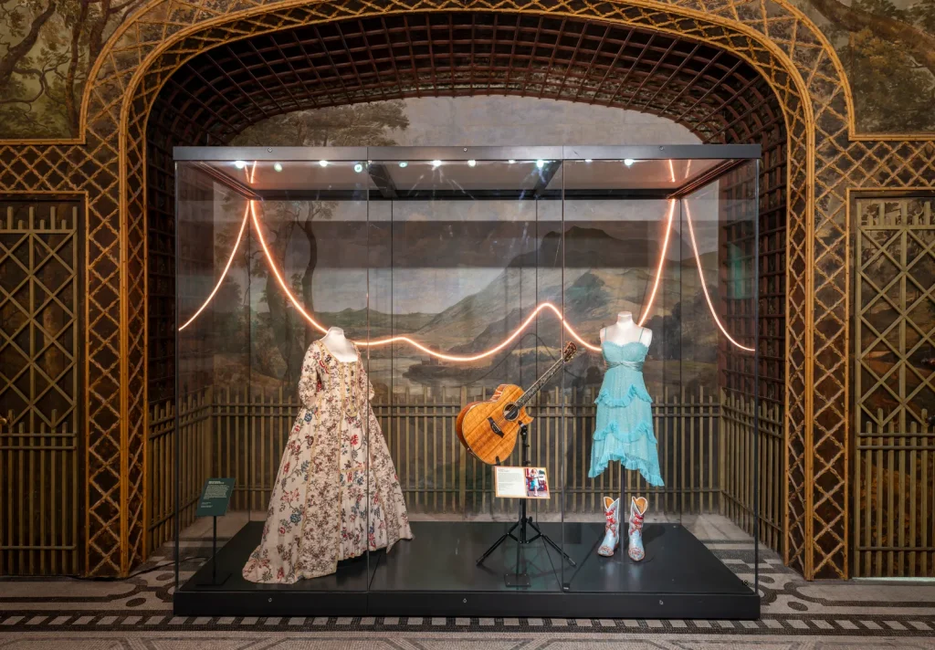 Two female mannequins ; ( Left) A historical dress, floor-length floral patterns and embellishments. (Right) A modern teal blue short dress with silver and white boots. Vitrine features a guitar.