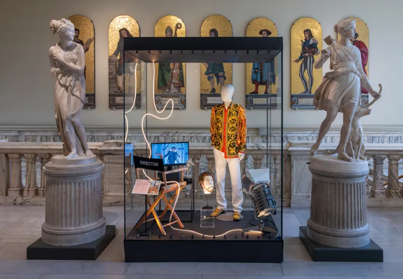 A male mannequin wearing a gold and orange shirt paired with cream trousers. The vitrine features a laptop, Director's chair, brown wig on wig stand and other 'film' related objects.