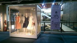 Three female mannequins (Left) wearing a short peach dress,(middle) wearing a short navy dress and (right) wearing a white wedding dress. Vitrine also displays a bouquet and white high heel shoes.