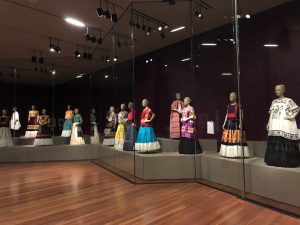 Fourteen female mannequins on display all wearing long tops with flowy skirts and dresses. Clothing are in hues of bright blue, purple, orange, red, green and black and white.