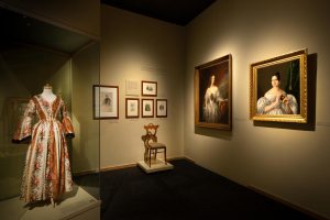 A floral historical dress on display complete with white and brown sleeves. Also on display are historical paintings of women.