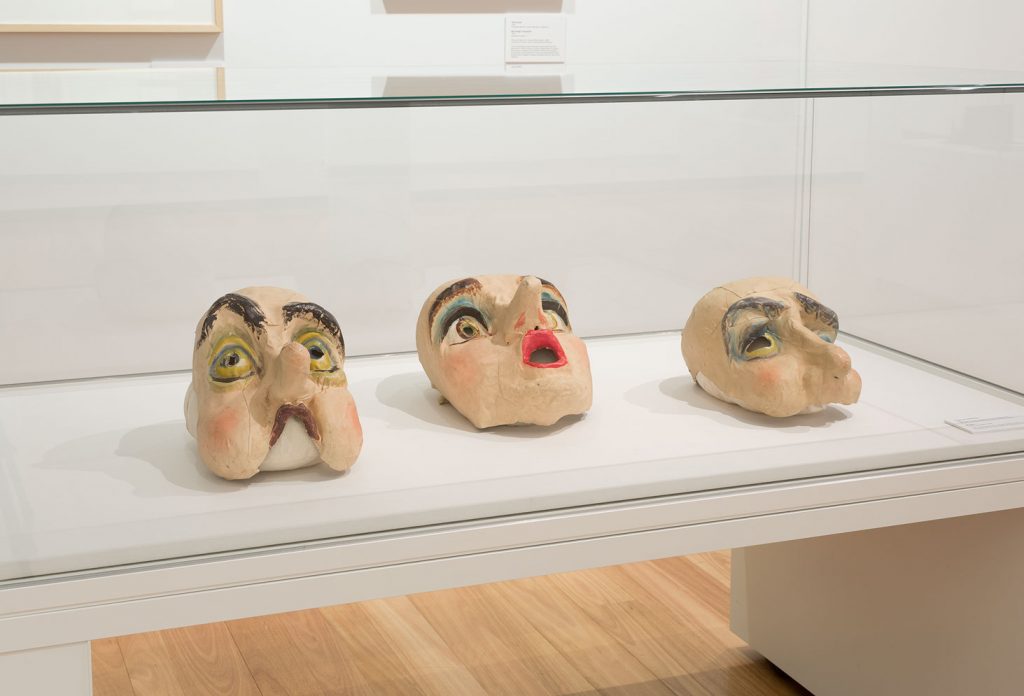 Three masks all expressing different emotions. Make up is in hues of peach, green, brown and blue.