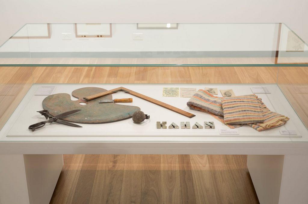 A vitrine displaying scissors black and grey that are open, an artist's painting palette, brown ruler, and pieces of cream and brown fabric.