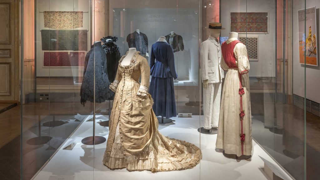 Eight mannequins on display in hues of dark blue, cream, red and gold, wearing men and women's eveningwear. Front two mannequins wearing a gold embellished full length dress and two piece red and white suit.