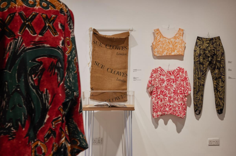 Five garments/ textile pieces on display. (Left) red and black kimono style outfit. (Left/middle) brown fabric hung from a white beam with black text printed on it. (Middle) yellow floral crop top paired with a red and white summer blouse. (Right) Yoga pants black and gold.