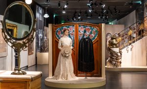 (Left) A white gown laced with floor length beige skirt, female mannequin is holding a brown straw hat. (Right) Half length black cloak.