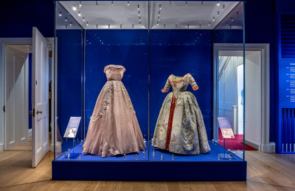 Two historical dresses on display. (Left) Pink ballgown dress with floral embellishments. (Right) Historical gown. blue with a red stripe in the centre of the gown.