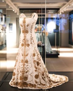 This is a floor-length, sleeveless gown with a fitted bodice and wide straps, featuring a soft ivory or off-white base colour. The dress is richly embellished with gold floral embroidery that cascades symmetrically down the front and continues onto the train, adding elegance and texture.
