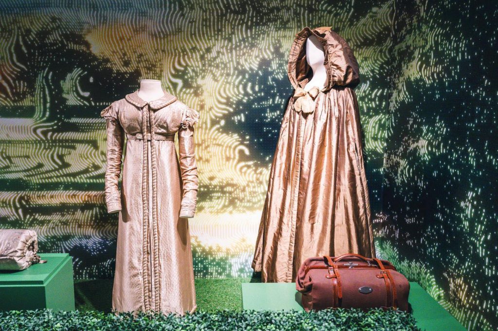 Two female mannequins on display (Left) Brown dress with a Peter pan collar and sleeves. (Right) hooded cloak brown and with brown bow.