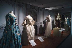 Four mannequins on display, (Left ) Teal blue gown with brown buttons on centre front of dress. (Left centre) A cream gown with long sleeves and ruffles. (Centre right) long white and puff sleeves. (Right) menswear on display, black jacket with gold trim complete with white shirt and trousers.