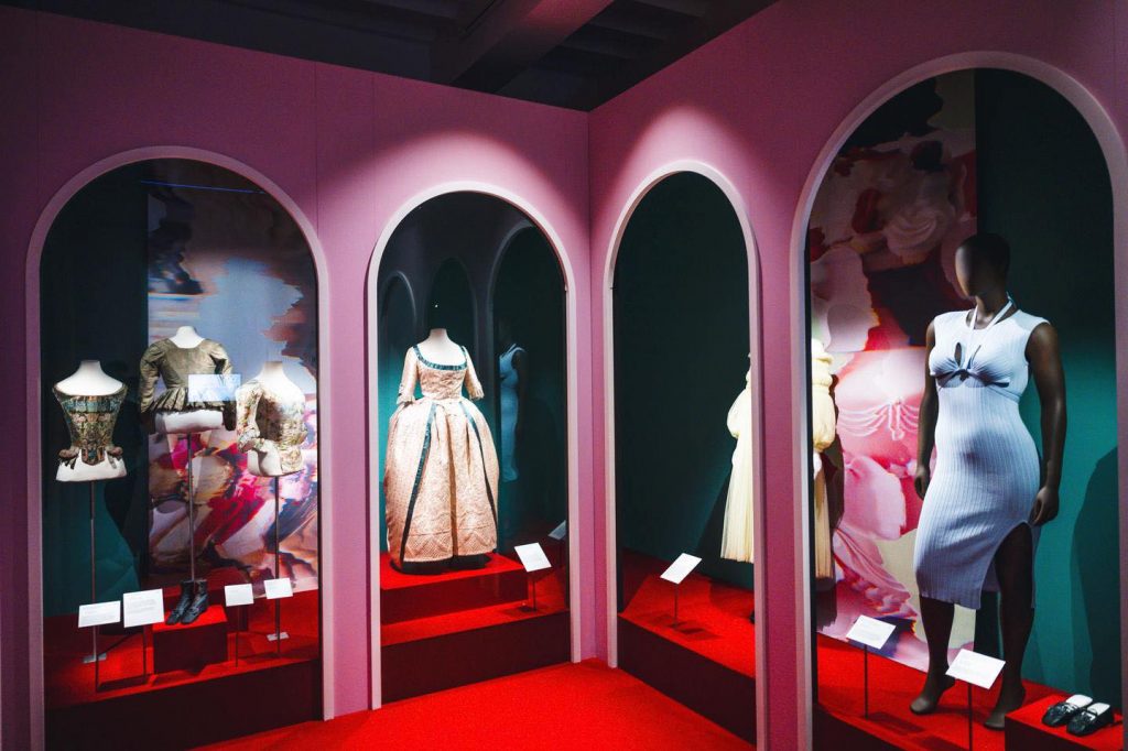 On the far left, there are three historical bodices in muted tones, olive green, beige, and floral patterns, accompanied by black shoes. The centre-left mannequin displays a full-length, formal gown in soft peach with teal accents, featuring a wide skirt and elbow-length sleeves, typical of the 18th-century Rococo style. The middle alcove showcases a white ribbed knit dress with a bodycon fit, keyhole cutouts near the chest, and a side slit—this piece is modern and minimalist. Behind that, a pale yellow or cream pleated dress with voluminous sleeves is partially visible, suggesting a romantic or avant-garde design. On the far right, a mannequin in a fitted sleeveless white dress emphasises a contemporary silhouette, standing beside a pair of black flat shoes.