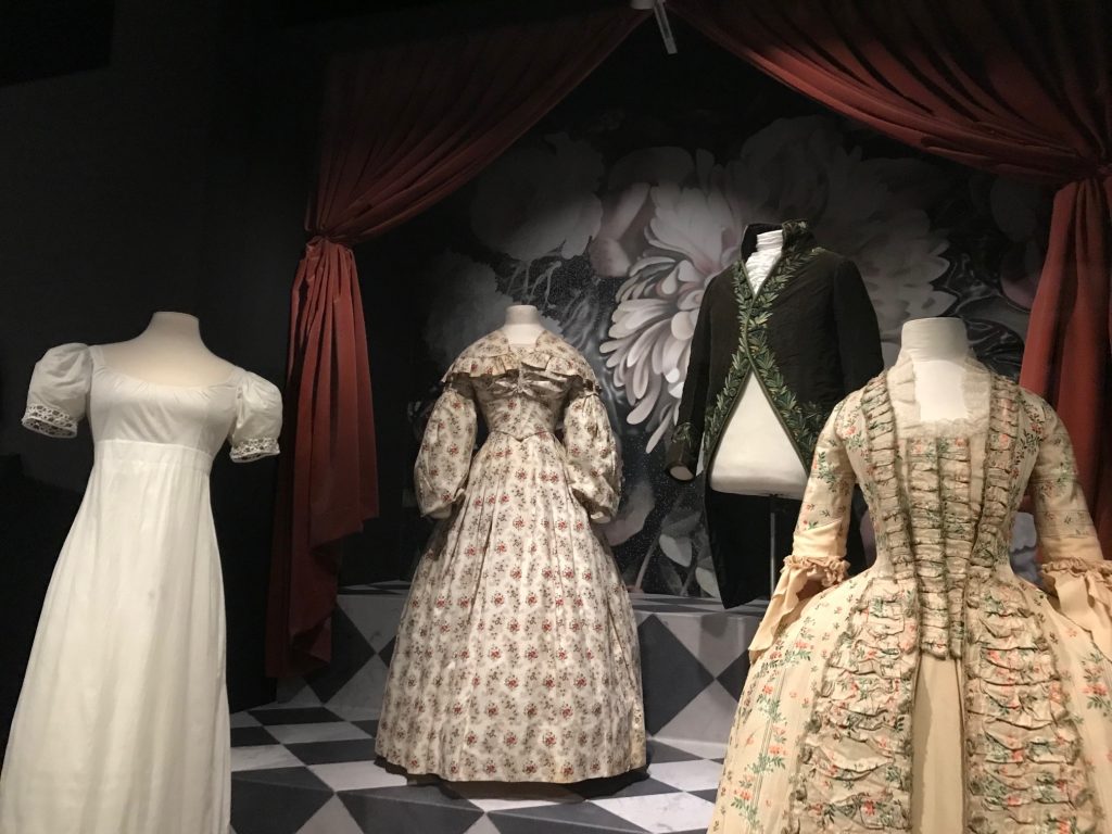 Four mannequins three females and one male. (Left) Female mannequin wearing a white 'chemise' like dress with puff sleeves. (Left centre) Long sleeved gown with floral pattern in cream. (Right centre) male black jacket with tails. (Right) cream with green piping with ruffles and sleeves.