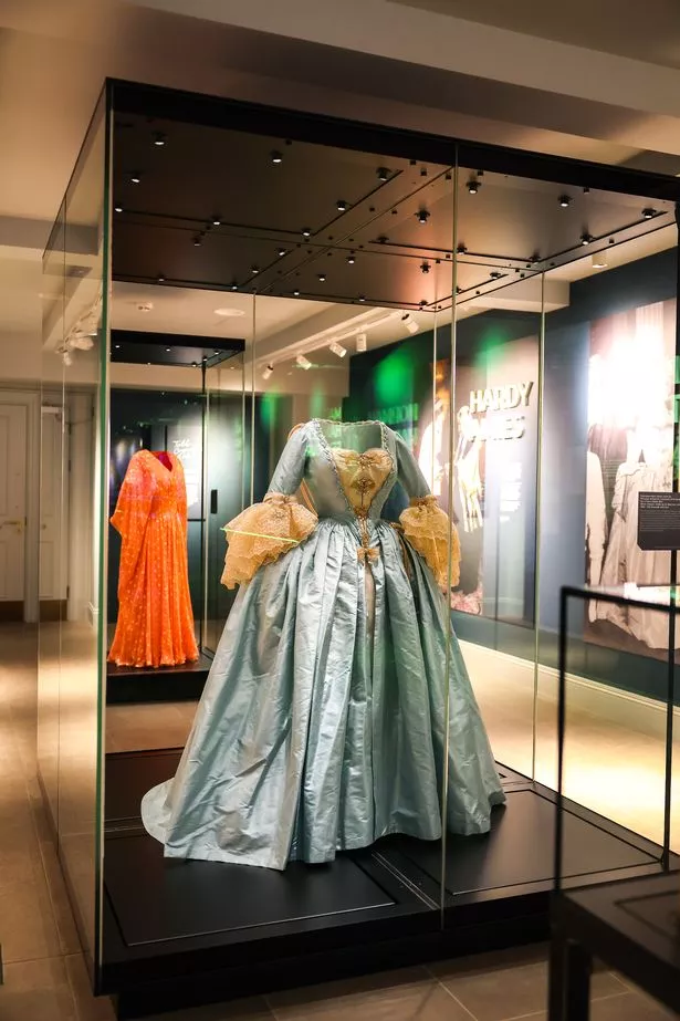 Pale blue ball gown with an off-the-shoulder neckline and a voluminous skirt, designed in a classic, mid-20th-century style. The bodice appears structured and fitted, while the full skirt creates an elegant, regal silhouette. Delicate embellishments like embroidery or beading can be faintly seen.