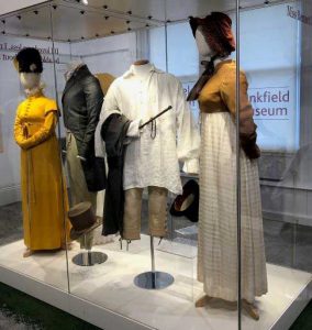 Four mannequins wearing typical regency fashion. (Left) Female mannequin wears a yellow dress with sleeves and a black hat. (Middle) male mannequin wearing a black jacket with tails. (Centre Right) A male white shirt with brown trousers. (Right) Female mannequin wearing a brown bonnet, a mustard yellow top with a white full length skirt.