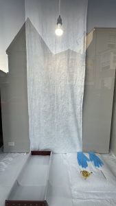 An installation inside a window front. The photograph shows the background of the window with a lightbulb that is switched on and a white sheet hanging in the centre. (Centre) A pair of blue nitrile gloves is laid neatly on a white cloth beside a golden accessory, possibly a brooch or decorative trim. To the right, an open archival storage box lined with tissue paper is ready for use.