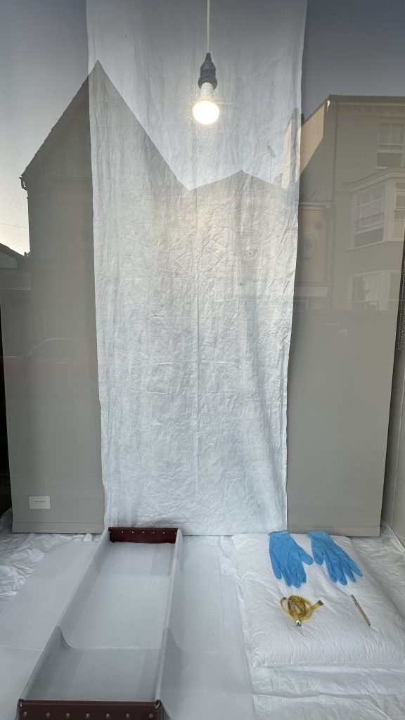 An installation inside a window front. The photograph shows the background of the window with a lightbulb that is switched on and a white sheet hanging in the centre. (Centre) A pair of blue nitrile gloves is laid neatly on a white cloth beside a golden accessory, possibly a brooch or decorative trim. To the right, an open archival storage box lined with tissue paper is ready for use.