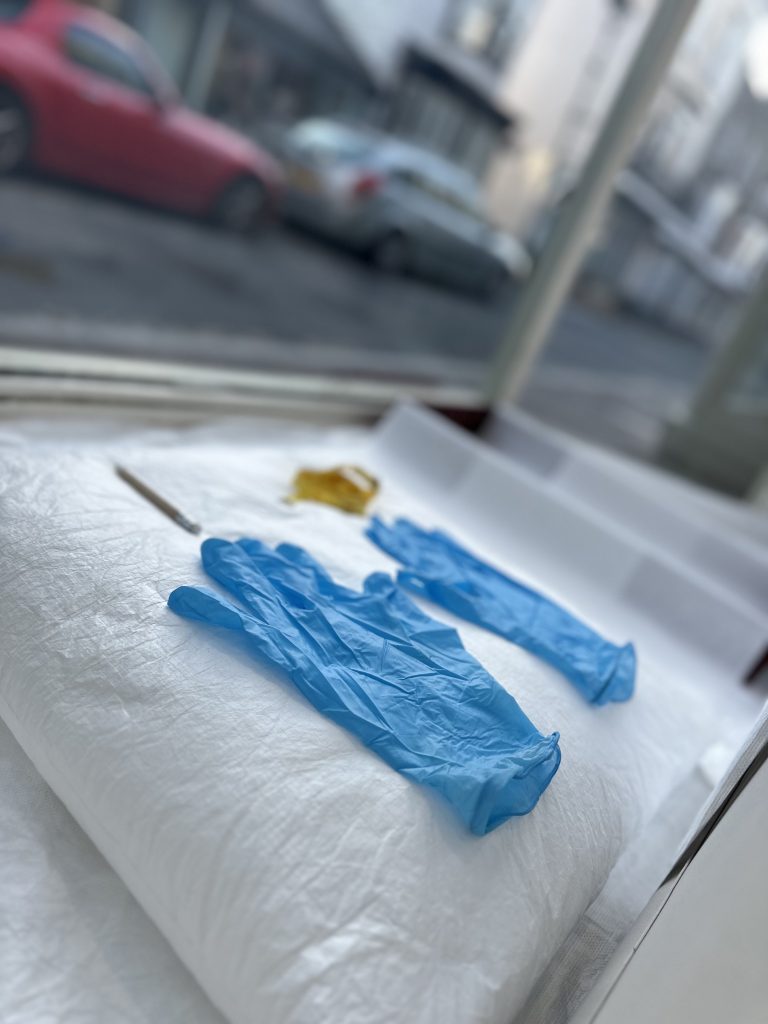 An installation inside a window front. Photograph is taken from a side view. A pair of blue nitrile gloves is laid neatly on a white cloth beside a golden accessory, possibly a brooch or decorative trim. To the right, an open archival storage box lined with tissue paper is ready for use.