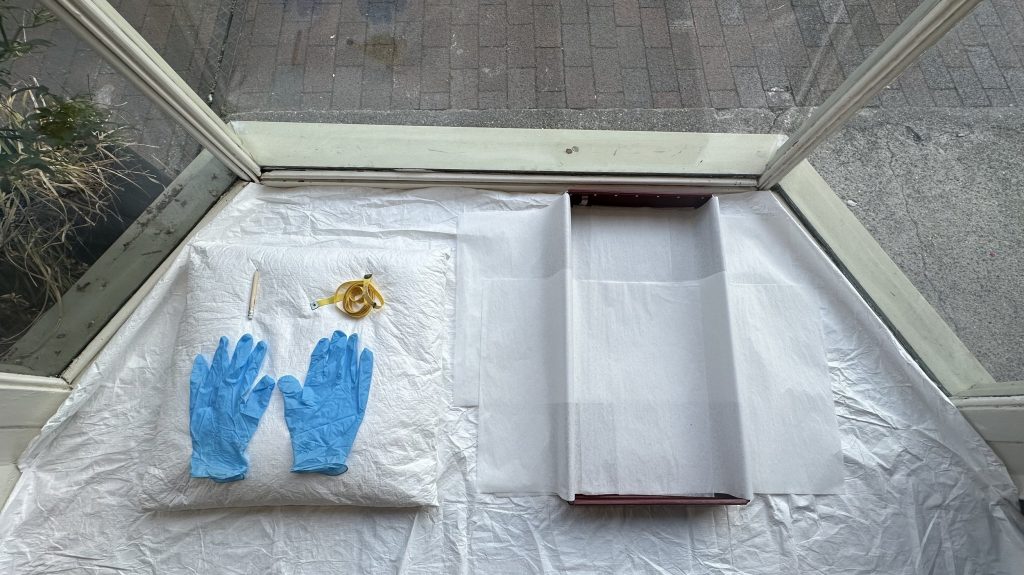 An installation inside a window front. (Centre) A pair of blue nitrile gloves is laid neatly on a white cloth beside a golden accessory, possibly a brooch or decorative trim. To the right, an open archival storage box lined with tissue paper is ready for use.