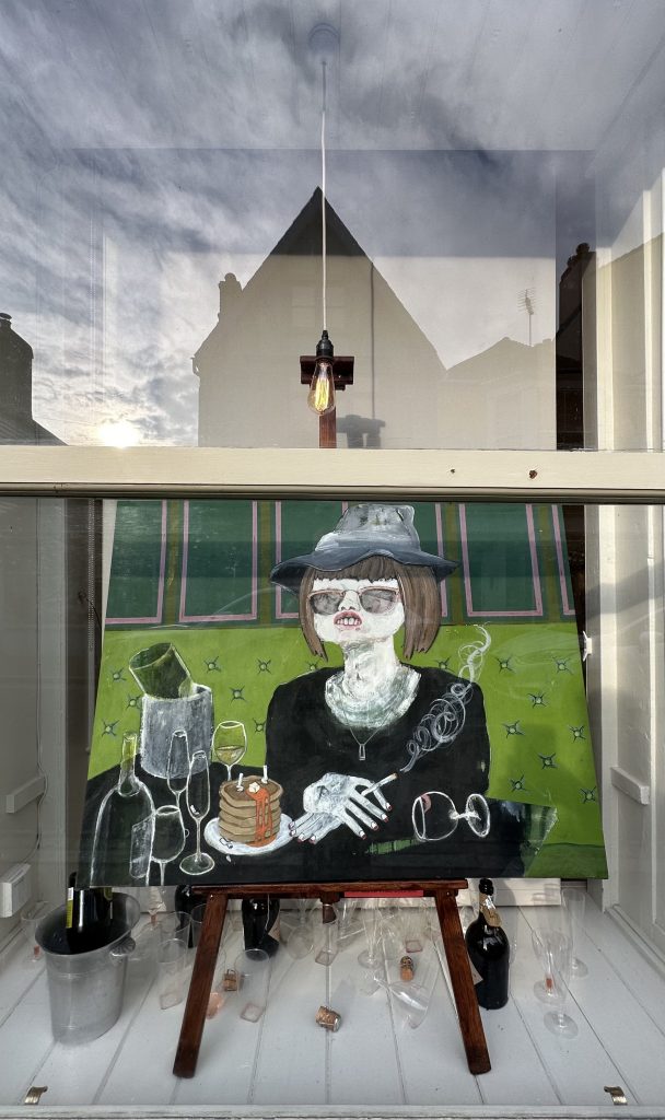 On display in the window is a vibrant painting of a female figure wearing a grey hat, glasses, black top and is smoking. The figure is posed at a black table with empty clear wine glasses, and food. On the floor are various empty wine glasses and a grey champagne bucket.