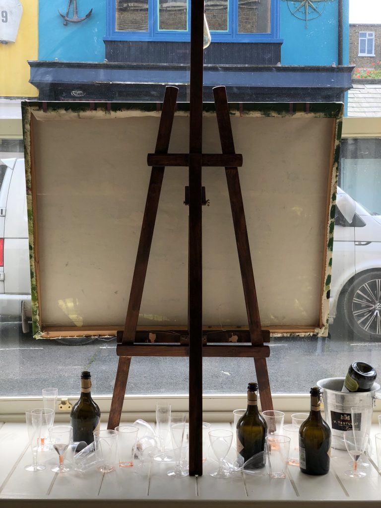 One window display is a large blank canvas is mounted backward on a wooden brown easel, facing the street. Scattered below are empty black and gold wine bottles, used clear glasses, and a tilted grey champagne bucket.