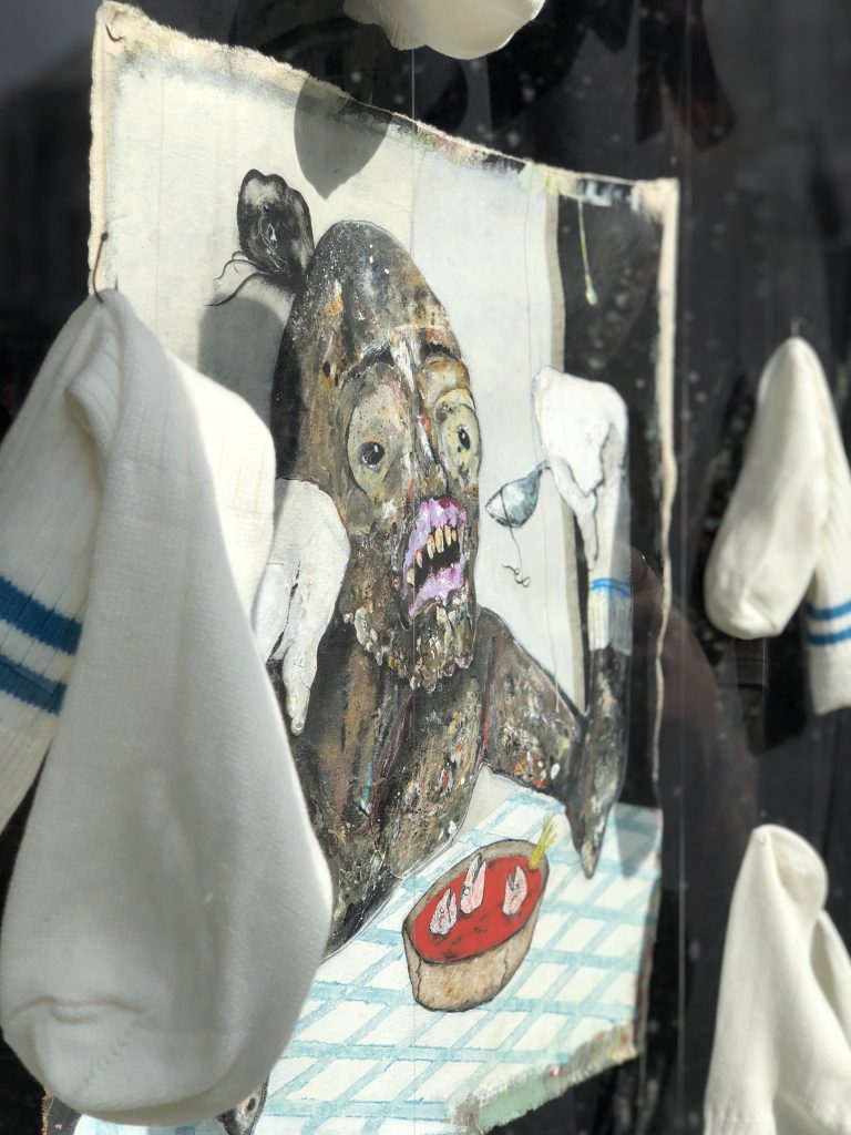 A close up of a figure in hues of grey, pink, red and green. The figure poises with a silver spoon and in front of the figure is a round dish with a red liquid inside. The painting is surrounded by suspended white socks with blue stripes.